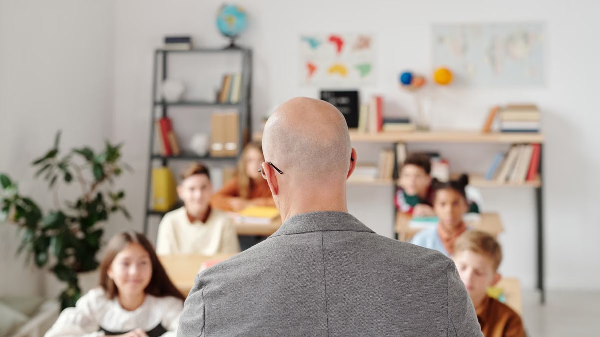 Bald teacher in a classroom engaging with attentive students sitting at desks.